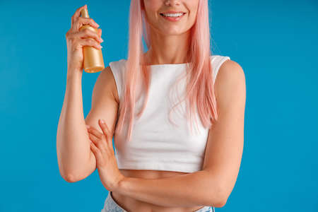 Cropped shot of young woman with pink hair smiling and holding bottle of moisturizing spray while...
