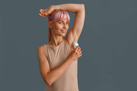 Happy young woman with pink hair in beige bodysuit smiling at camera while using antiperspirant, posing isolated over gray studio backgroundの写真素材