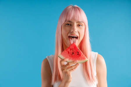 Playful young woman with pink hair holding a slice of watermelon, posing with mouth open isolated over blue studio backgroundの写真素材