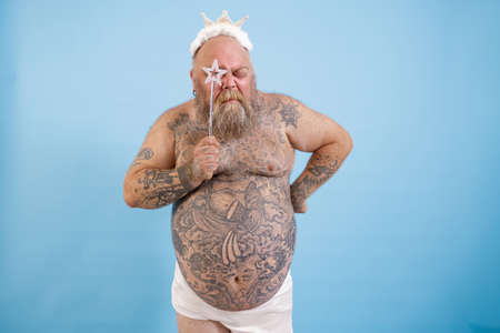 Tattooed obese man with crown looks through star on magic stick on blue backgroundの写真素材