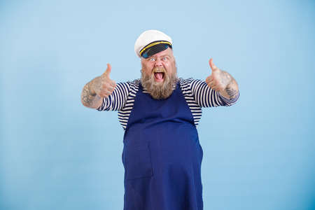 Emotional male person with overweight in sailor suit a shows thumb up on light blue background