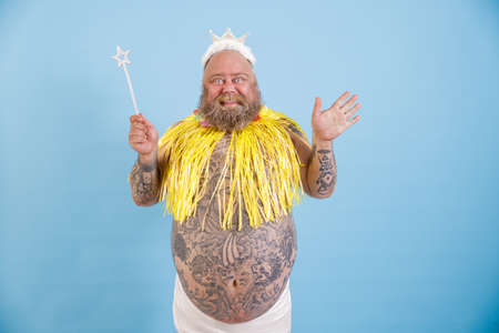 Joyful bearded plump man with crown, magic stick and yellow cape on light blue backgroundの写真素材