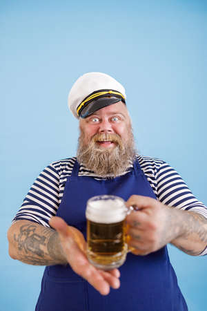 Cheerful plump man in sailor costume offers glass mug of beer on light blue backgroundの写真素材