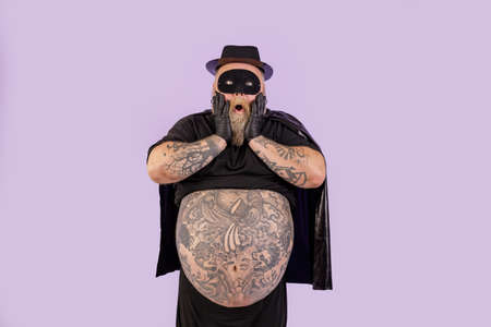 Shocked plump man in hero costume holds cheeks standing on purple backgroundの写真素材