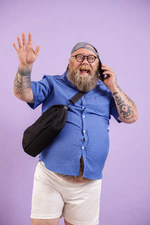 Jouful plump man talks by phone and waves hand standing on purple backgroundの写真素材