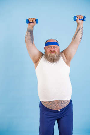 Fat man does sports exercises with dumbbells trying to loss weight on light blue backgroundの写真素材