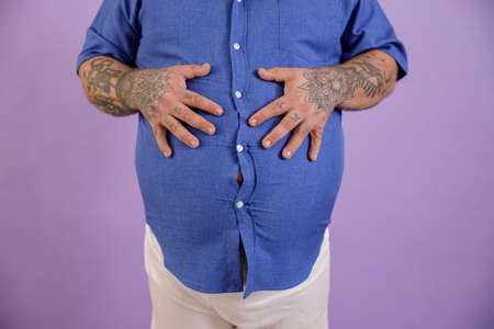 Man with overweight and tattoos holds hands on large belly on purple backgroundの写真素材