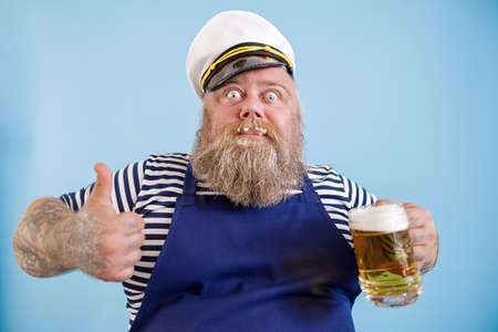 Joyful fat person in sailor suit holds beer and shows thumb up on light blue backgroundの写真素材