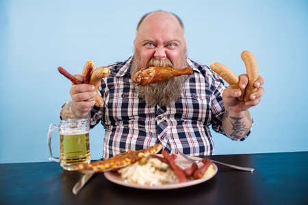 Fat man with chicken leg in mouth holds sausages at table in studioの写真素材