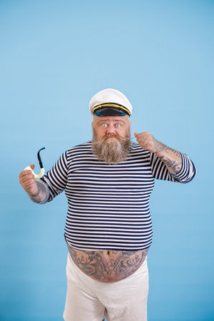 Bearded obese man sailor with smoking pipe tightens up mustache on light blue backgroundの写真素材