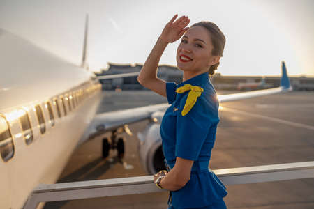 Portrait of cheerful air stewardess in blue uniform smiling at camera, posing outdoors at the sunset. Commercial airplane near the terminal in an airport in the backgroundの写真素材
