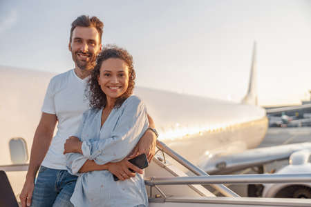 Portrait of happy couple of tourists, man and woman looking excited while standing together outdoors ready for boarding the plane at sunsetの写真素材