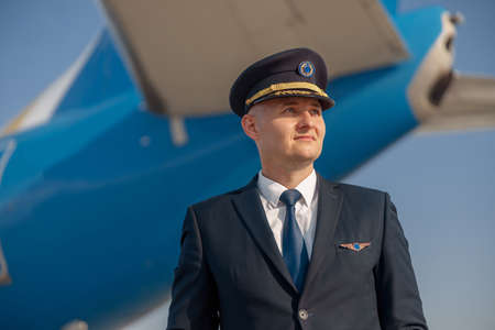 Portrait of pilot in uniform looking away, standing in front of big passenger airplane ready for departure in airportの写真素材