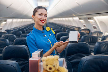Pretty stewardess in bright blue uniform smiling at camera, advertising perfume as a gift while leading trolley cart through empty plane aisleの写真素材