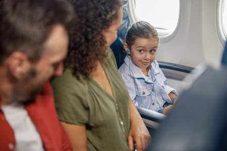 Cute little girl wearing earphones while sitting on the plane, listening to music, looking at her parents, traveling together with familyの写真素材