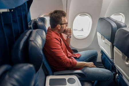Pensive caucasian man in casual wear resting on the plane, sitting and looking out the window