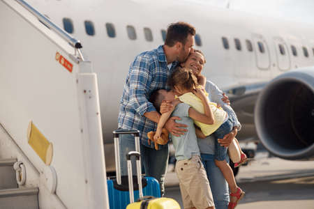 Family of four kissing each other while going on a trip, standing in front of big airplane outdoorsの写真素材