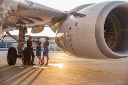 Happy pilot and two pretty stewardesses standing together, leaning on an airplane and smiling at camera after landing or before departureの写真素材