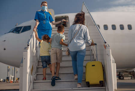 Back view of mother with two little kids and suitcase boarding the plane. Air stewardess wearing protective mask welcoming the familyの写真素材