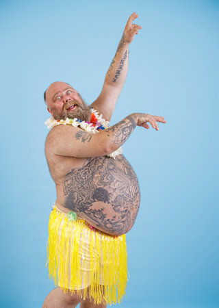 Funny obese man in decorative grass skirt and garland dances on light blue backgroundの写真素材