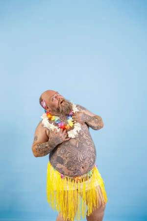 Surprised plump man in yellow grass skirt and garland looks up on light blue backgroundの写真素材