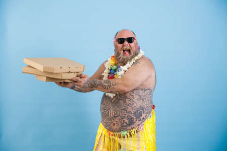 Happy obese man in grass skirt and flowers garland holds pizzas on light blue backgroundの写真素材