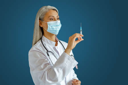 Middle aged doctor with medical mask on blue background, focus on hand with syringeの写真素材