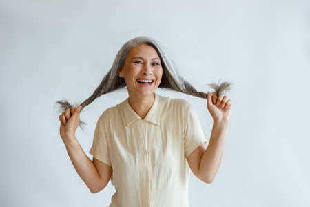 Happy middle aged Asian woman holds twisted hoary hair on light grey backgroundの写真素材