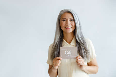 Smiling grey haired Asian woman holds gift card standing on light background