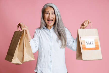 Excited mature Asian woman holds paper bags with colorful Sale sign standing in studioの写真素材