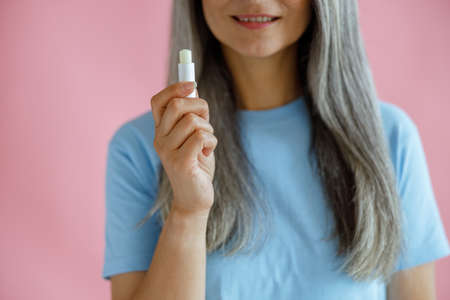 Mature grey haired woman on pink background closeup, focus on hand with lip balmの写真素材