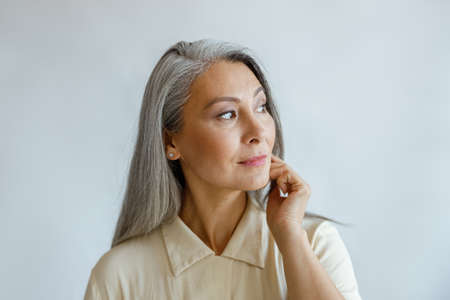Thoughtful grey haired woman in stylish shirt looks aside on light backgroundの写真素材