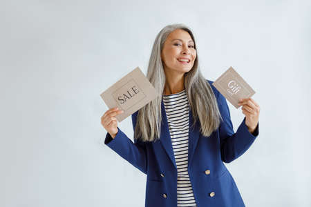 Cheerful middle aged Asian woman holds gift card and Sale sign standing in studioの写真素材