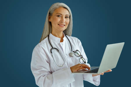 Smiling mature woman doctor in white coat works on laptop standing on blue backgroundの写真素材