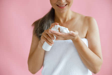 Smiling middle aged lady with towel squeezes body care product onto hand on pink backgroundの写真素材