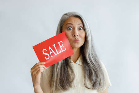 Funny middle aged Asian woman shows red Sale sign grimacing on light backgroundの写真素材