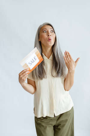 Emotional hoary haired Asian woman holds colorful card with word Sale on light background