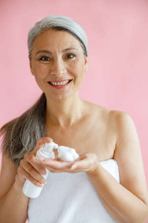 Positive middle aged Asian lady wrapped with soft towel holds cleansing foam on pink backgroundの写真素材