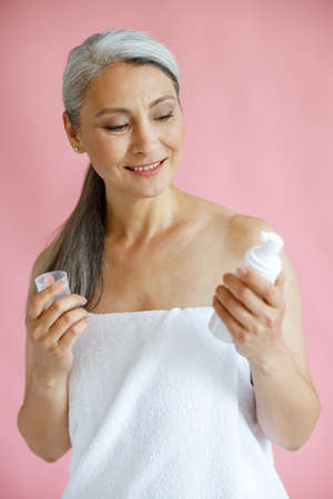 Pretty grey haired Asian woman looks at body care product on pink backgroundの写真素材