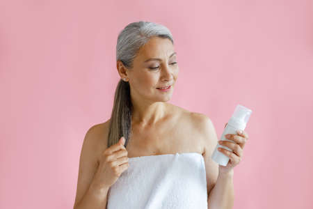 Beautiful mature Asian lady wrapped with towel holds bottle of body care product in studioの写真素材