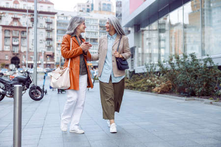Cheerful senior woman with long haired companion walk on large city street on autumn dayerの写真素材
