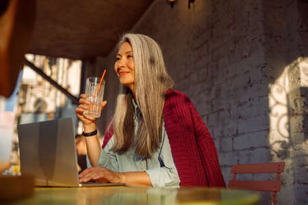 Dreamful senior Asian lady holds glass sitting at small table with open laptop