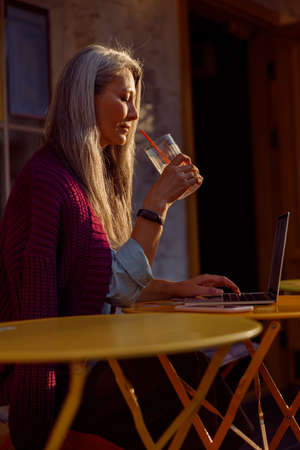 Senior Asian woman holds glass sitting in front of laptop at table on cafe terrace