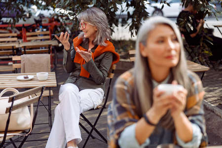 Senior cafe guests, focus on silver haired woman with glasses recording audio messageの写真素材