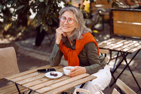 Positive senior woman with glasses and cup of coffee at table on outdoors cafe terraceの写真素材