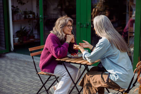 Silver haired woman tells story to mature friend sitting at table outdoorsの写真素材