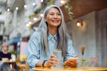 Cheerful mature Asian woman with glass of water and phone sits at small table outdoorsの写真素材