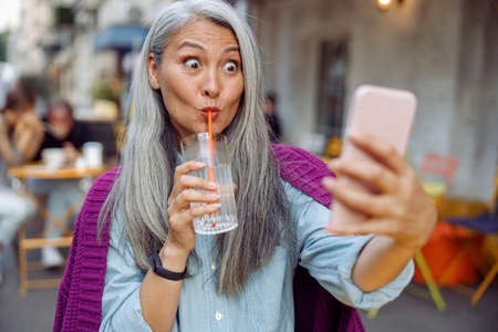 Funny mature Asian woman drinks water taking selfie with cellphone in cafe