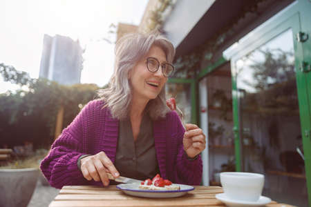 Joyful senior lady in purple jacket eats dessert with strawberries sitting at table outdoorsの写真素材