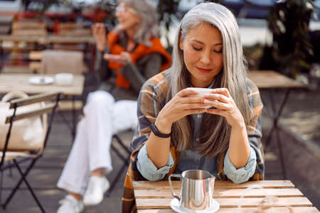 Senior cafe guests, focus on pretty silver haired lady holding cup of drinkの写真素材